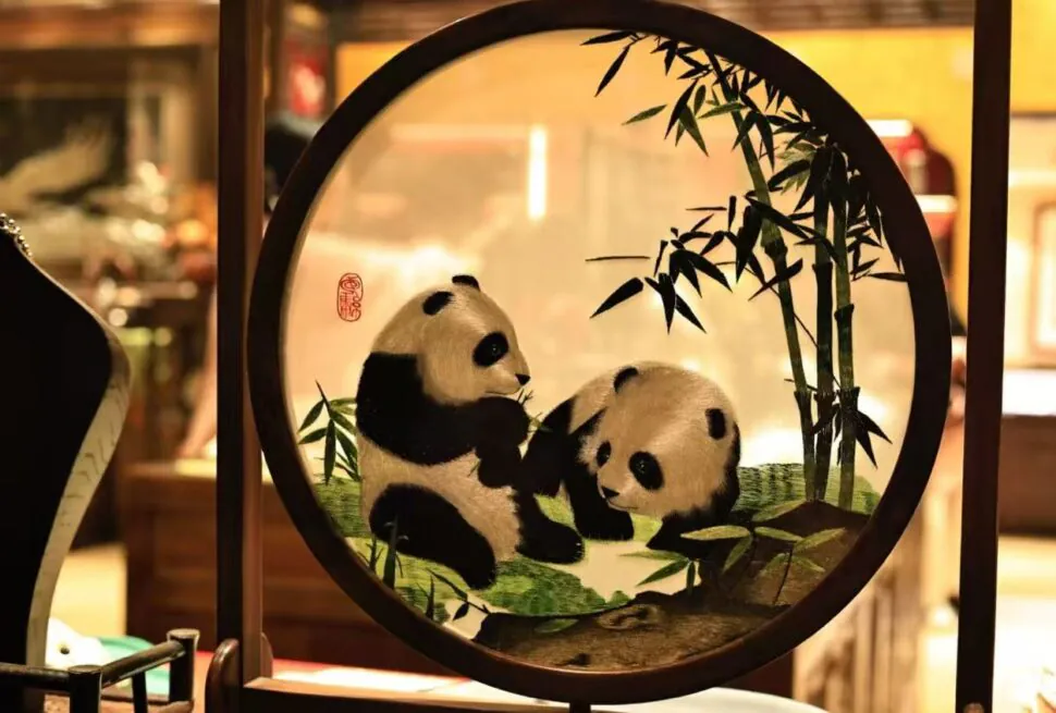 Two panda cubs playing with bamboo inside a circular decorative frame, an art piece on display in a warm shop setting.