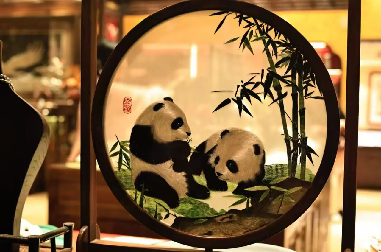 Two panda cubs playing with bamboo inside a circular decorative frame, an art piece on display in a warm shop setting.