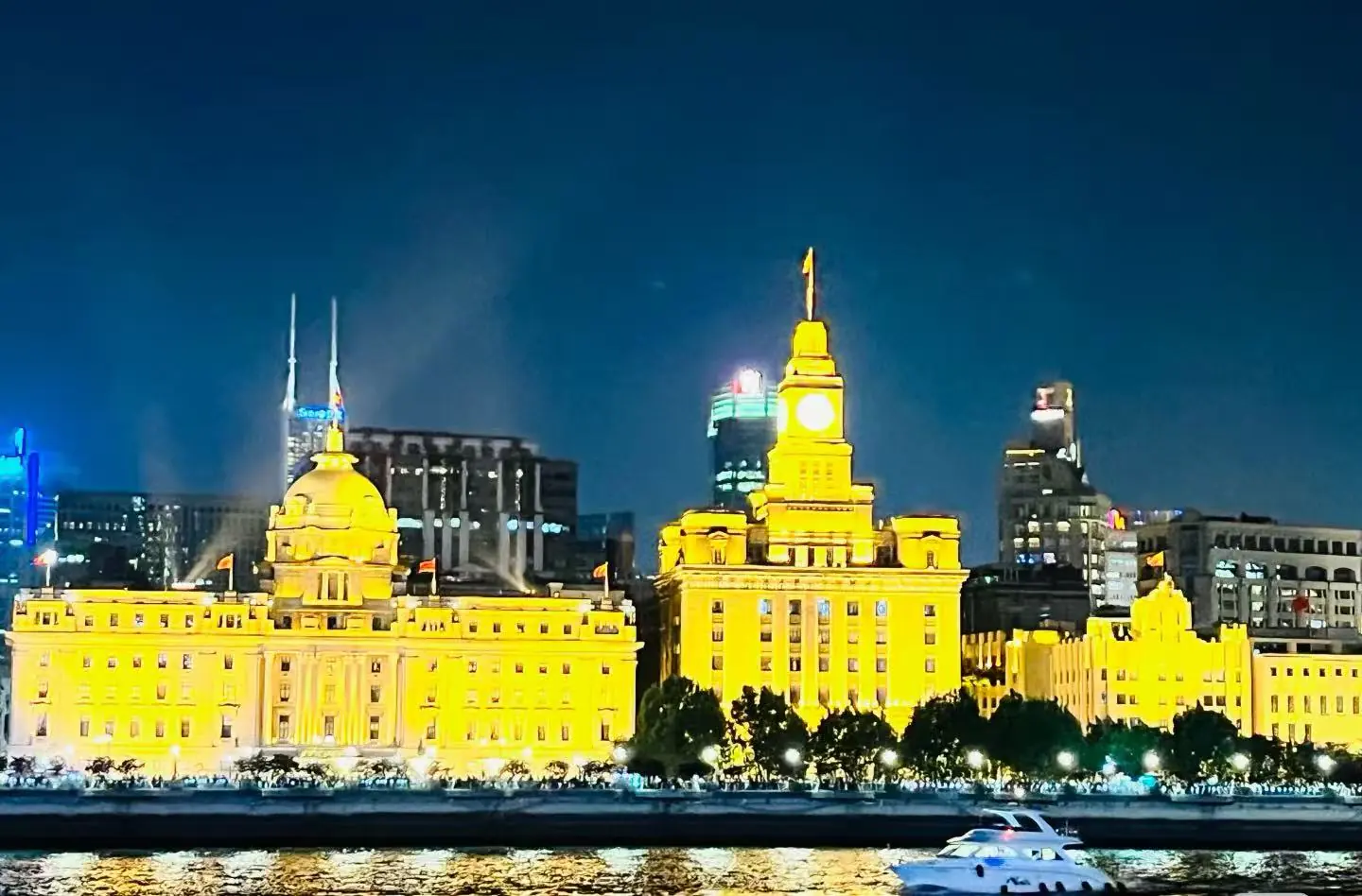 Nighttime cityscape with warmly lit, yellow historic buildings along a river and a white motorboat in the foreground.