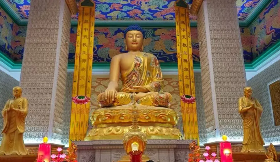 Famen Temple,X`an,Chinatour Golden seated Buddha statue in a richly decorated temple hall, flanked by two smaller statues, with a colorful ornate ceiling and illuminated altar candles and flowers behind it.