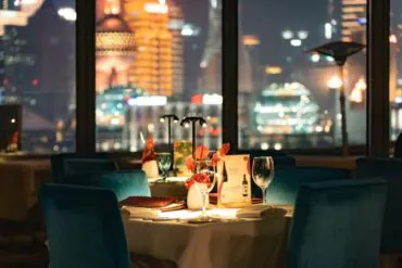 Table for two in a dim restaurant with teal chairs, a floral centerpiece, wine glass, and city lights outside the windows.