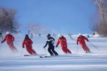 Group of skiers in red suits carving turns on a sunny snowy slope; a black-clad instructor leads.