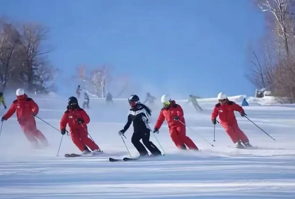 Group of skiers in red suits carving turns on a sunny snowy slope; a black-clad instructor leads.