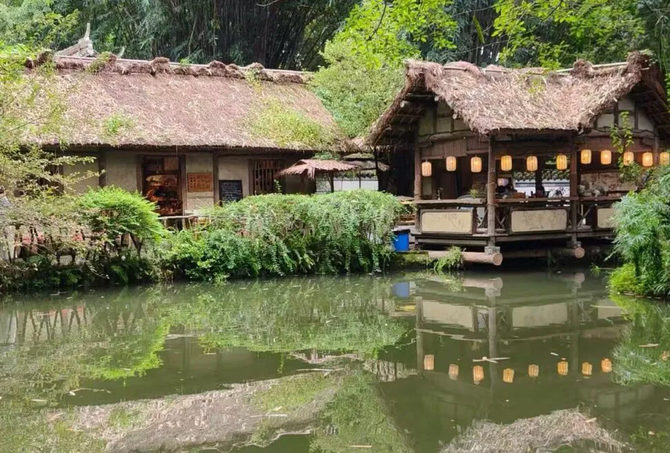 Du Fu Thatched Cottage,Chengdu,Chinatour Thatched-roof riverside cafe with lanterns, wooden veranda, and reflected greenery in a calm pond.