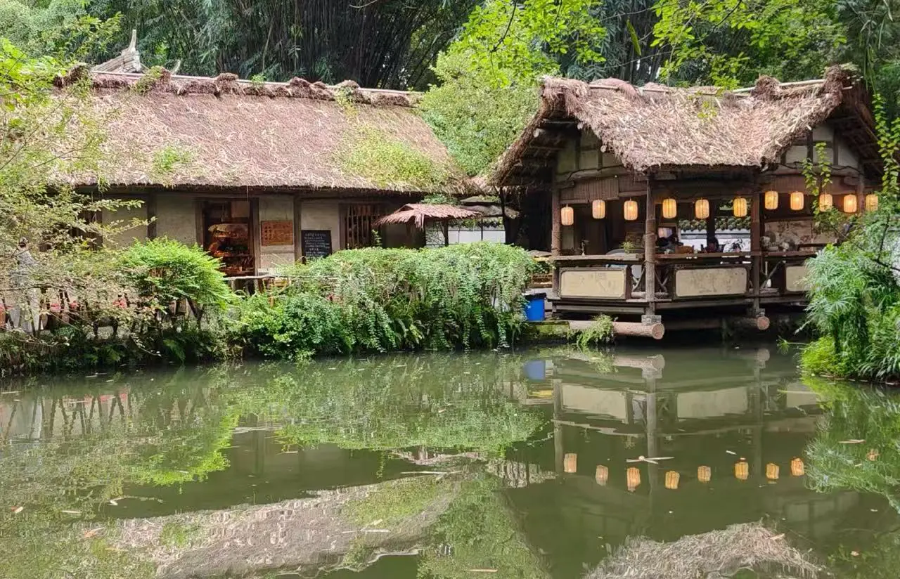 Du Fu Thatched Cottage,Chengdu,Chinatour Thatched-roof riverside cafe with lanterns, wooden veranda, and reflected greenery in a calm pond.