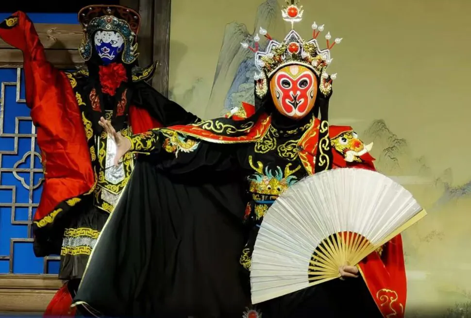 Two performers in ornate Chinese opera costumes with painted masks; one holds a white fan while the other strikes a pose in a red-and-black outfit.
