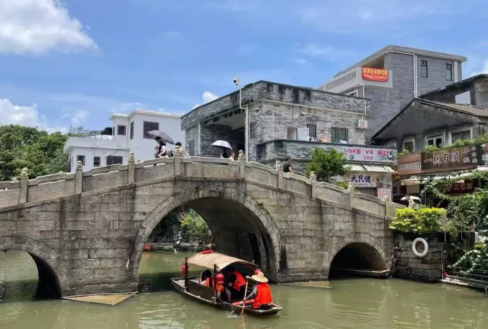 Stone arch bridge over a canal with pedestrians holding umbrellas on the bridge and a small tour boat passing beneath.