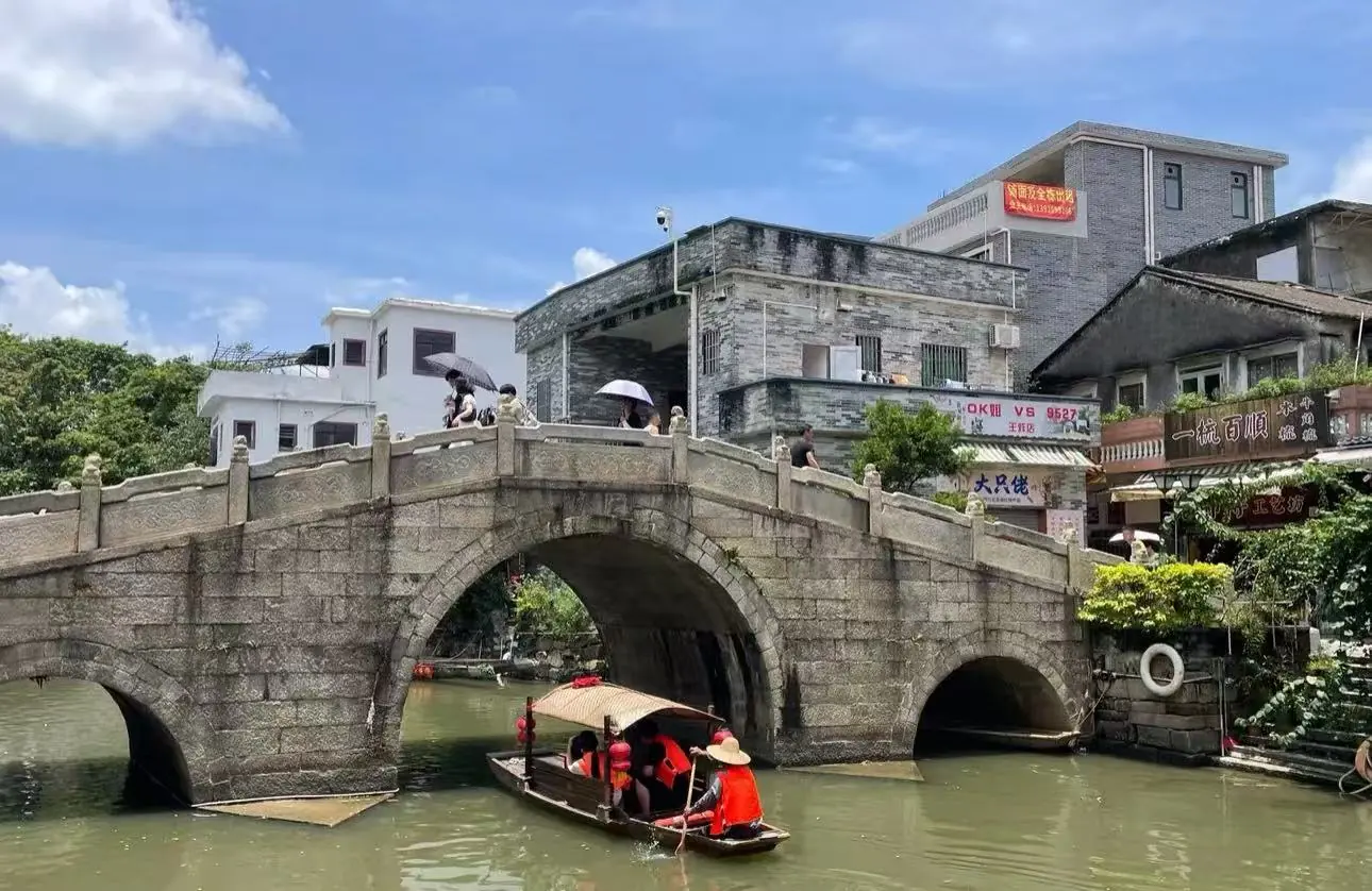Stone arch bridge over a canal with pedestrians holding umbrellas on the bridge and a small tour boat passing beneath.