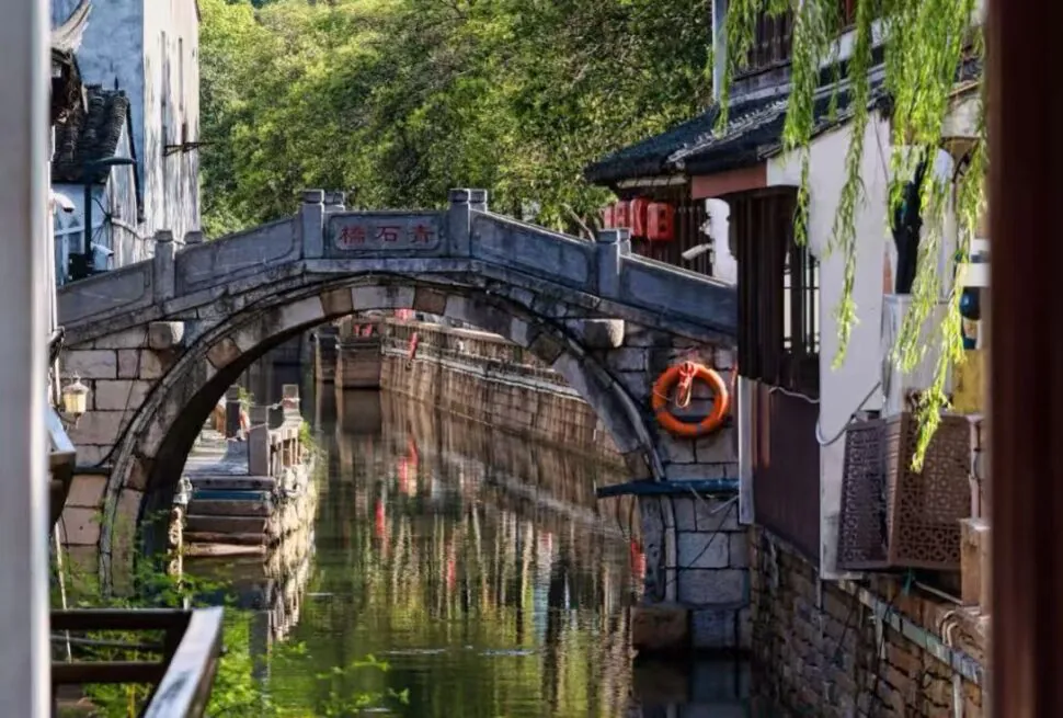 Stone arch bridge over a narrow canal in a traditional Chinese water town, with green trees and lanterns nearby.