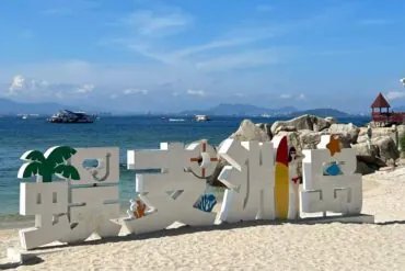 Large white three-dimensional decorative sign on a sandy beach by clear blue water, with palm-tree shapes and colorful accents, background rocks and boats on the sea.