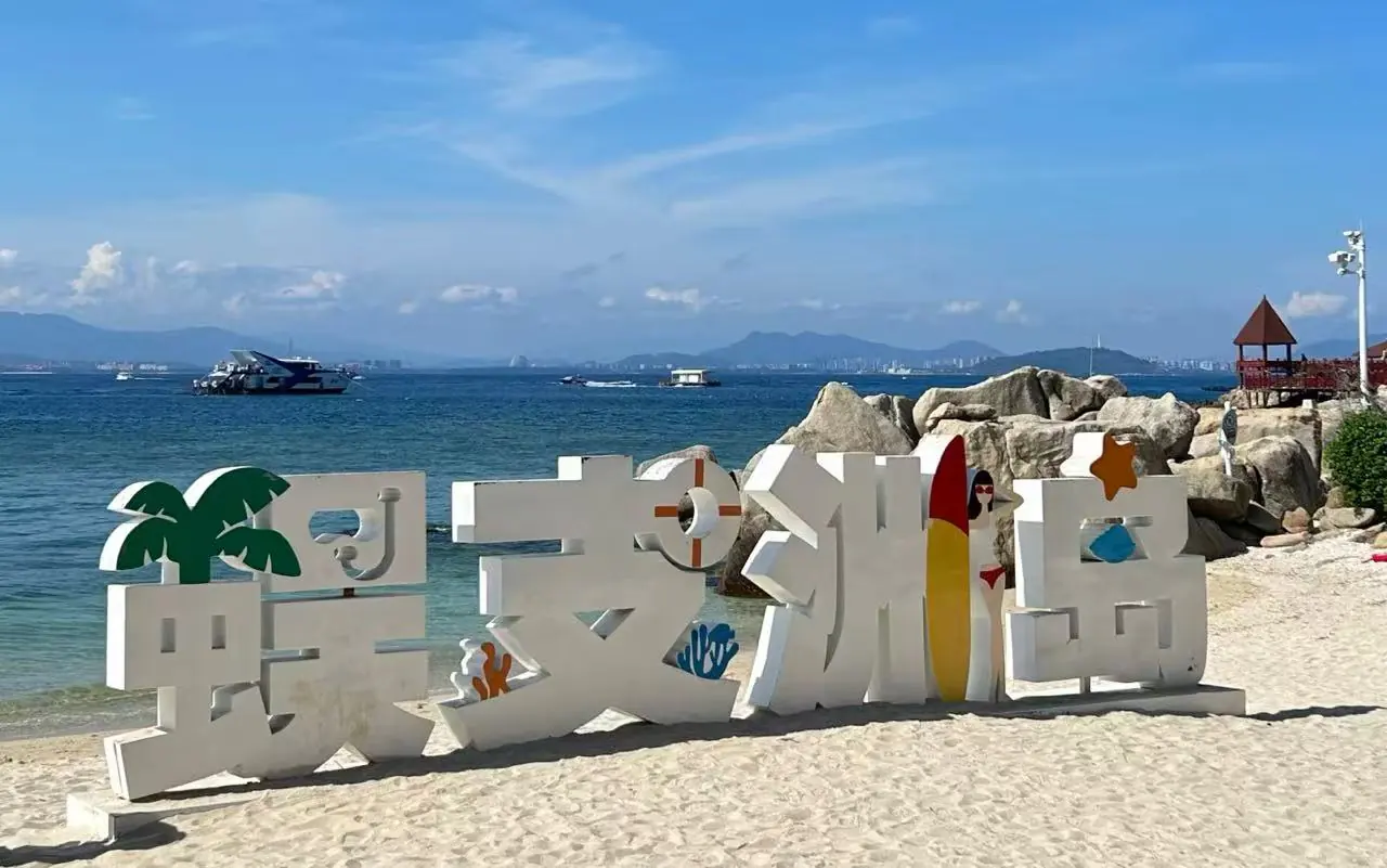 Large white three-dimensional decorative sign on a sandy beach by clear blue water, with palm-tree shapes and colorful accents, background rocks and boats on the sea.