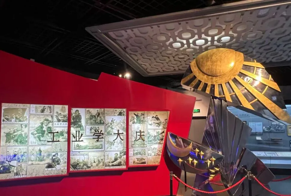 Daqing Oilfield History Museum,Daqing,Chinatour Museum exhibit with a red wall displaying comic panels and Chinese characters alongside a large metallic sunburst sculpture roped off in the foreground.