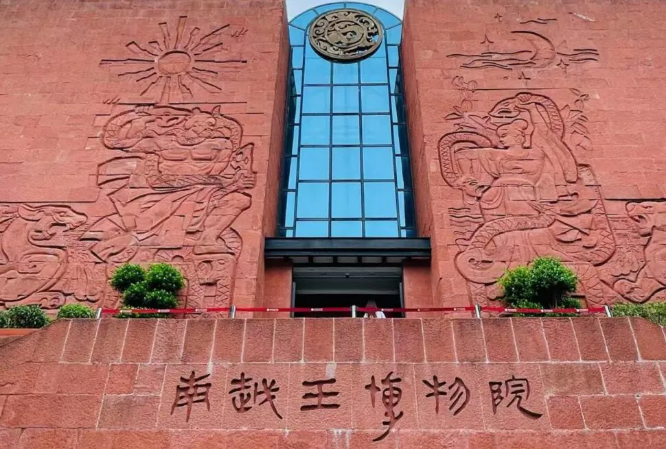Front view of a red brick museum facade featuring large stone reliefs on both sides, a blue glass central tower, and Chinese characters along the lower wall.