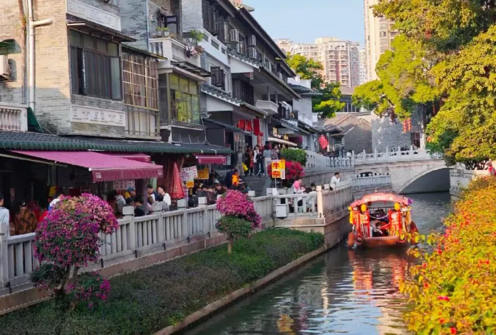 Crowded riverside street with shops and pink flowers along a canal, a colorful boat gliding by.