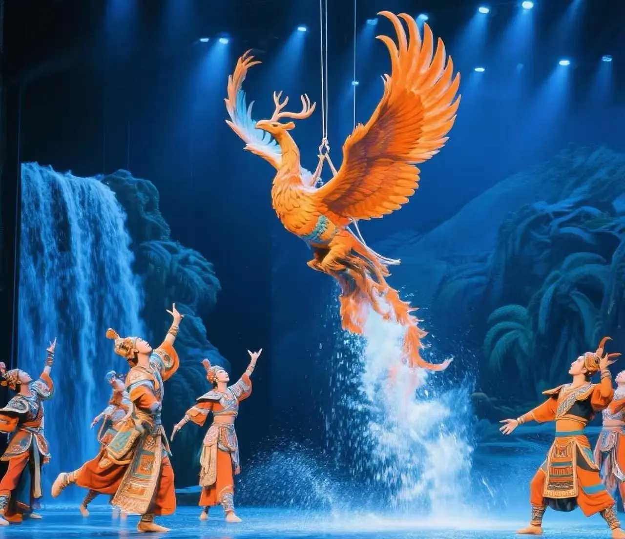 Sanya Romance Park Historical Shows,Sanya,Chinatour Large orange phoenix prop soaring on wires above dancers in colorful traditional costumes on a blue-lit stage with a waterfall backdrop.