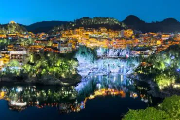 Night view of a hillside town with orange-lit buildings along a river, reflections in the water on a calm night.