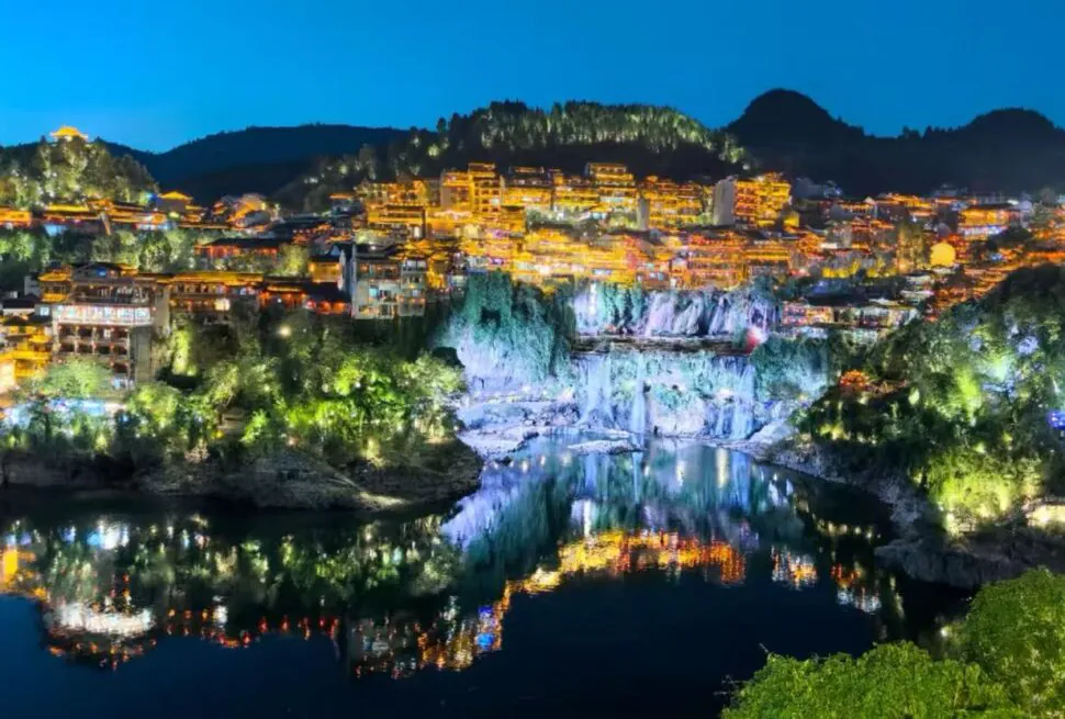 Night view of a hillside town with orange-lit buildings along a river, reflections in the water on a calm night.