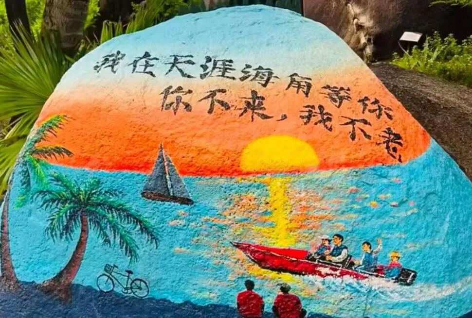 Tianya Haijiao Adventure,Sanya,Chinatour Colorful mural-painted rock showing sunset over the sea, palm trees, a sailboat, a red motorboat with people, two onlookers, a bicycle silhouette, and Chinese text above.
