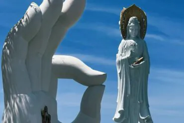 Giant white hand sculpture reaching toward a standing Bodhisattva statue with a halo against a clear blue sky.