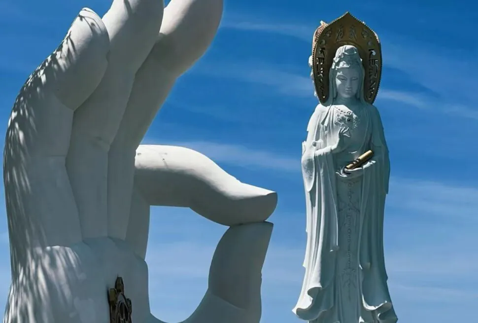 Nanshan Cultural Tourism Zone,Sanya,Chinatour Giant white hand sculpture reaching toward a standing Bodhisattva statue with a halo against a clear blue sky.