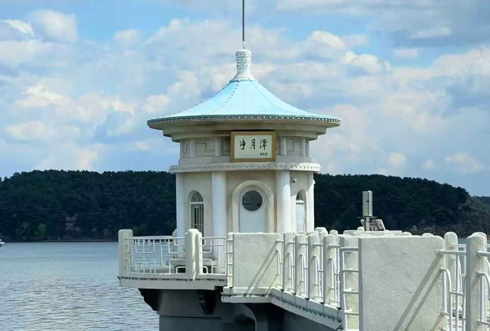 White round pavilion with a blue-teal roof on a pier over calm water, with trees in the distance.