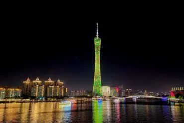 Guangzhou Canton Tower lit green at night beside a riverfront city skyline with reflections on the water, illuminated buildings in the background.