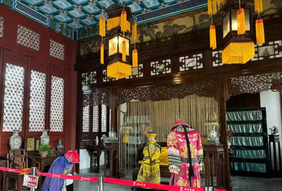 New Yuanming Palace,Zhuhai,Chinatour Inside a traditional Chinese hall with dark carved wood, gold-yellow lanterns, and red walls; two people in ornate dynastic costumes stand near a ceremonial altar.