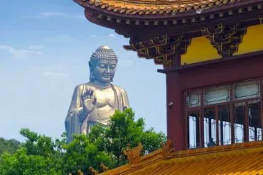 Large silver Buddha statue with right hand raised in blessing beside a yellow-orange ornate temple roof against a blue sky.