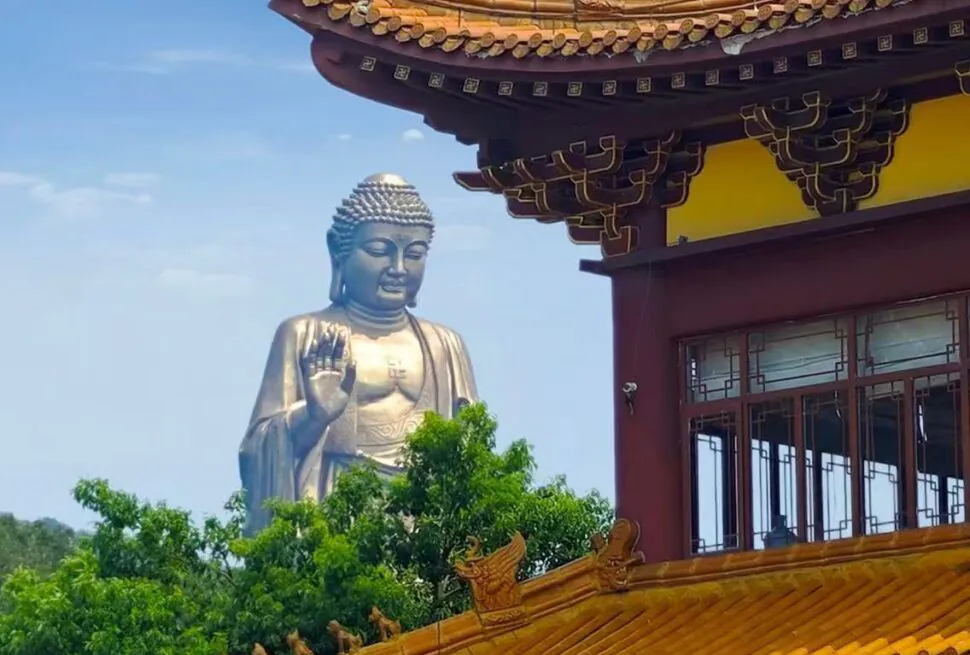 Large silver Buddha statue with right hand raised in blessing beside a yellow-orange ornate temple roof against a blue sky.