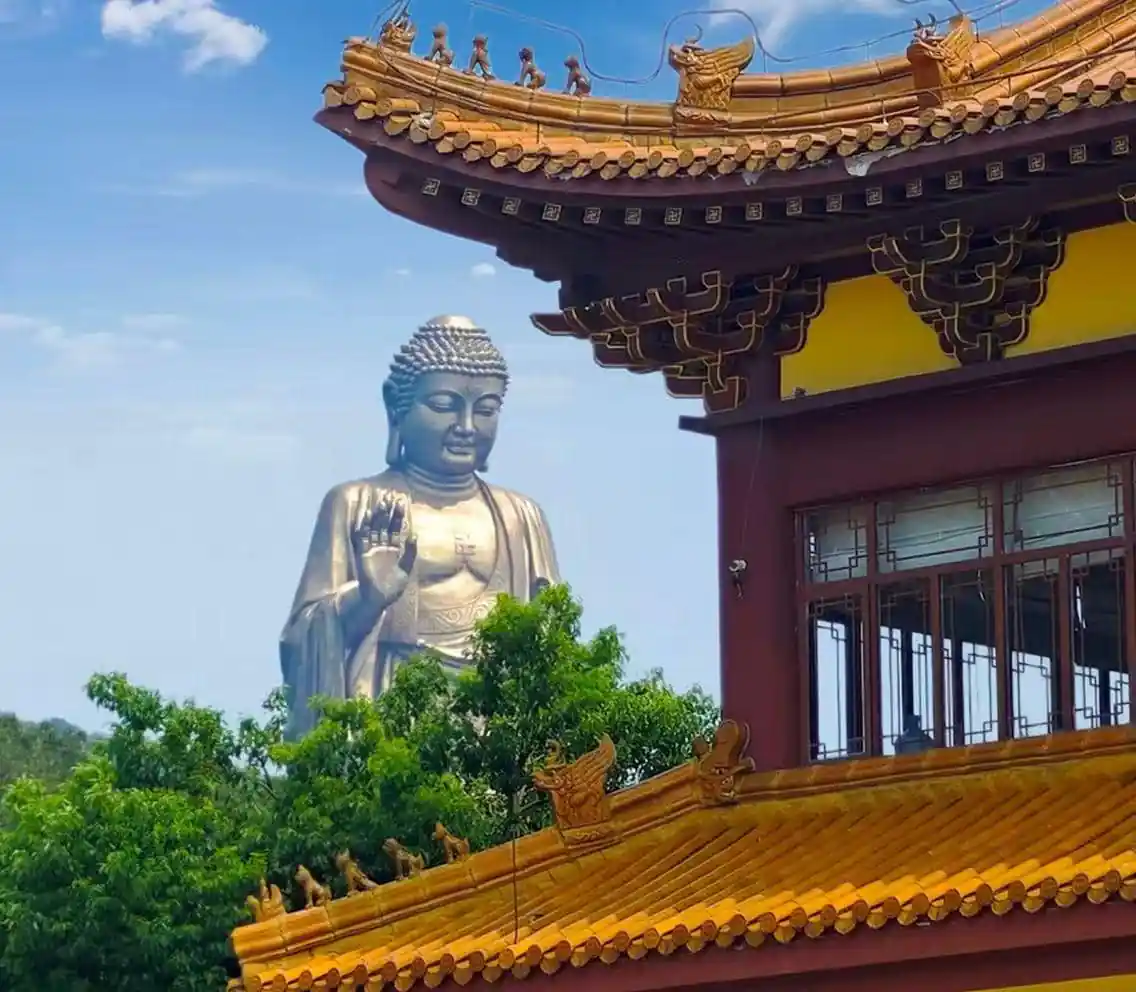 Large silver Buddha statue with right hand raised in blessing beside a yellow-orange ornate temple roof against a blue sky.