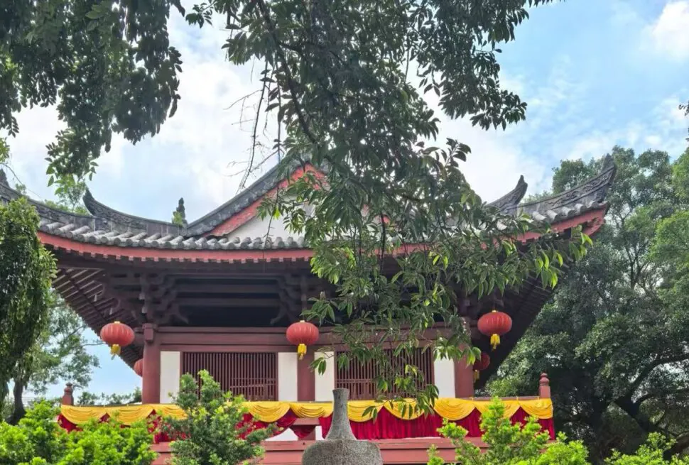 Traditional Chinese pavilion with curved tiled roof, red lanterns, and yellow drapery in a lush garden.