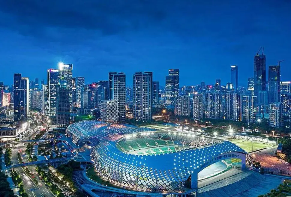 Aerial night view of a futuristic stadium with a blue lattice roof beside a dense illuminated city skyline.