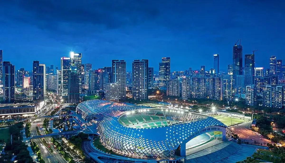 Shenzhen Bay Sports Center,Shenzhen,Chinatour Aerial night view of a futuristic stadium with a blue lattice roof beside a dense illuminated city skyline.