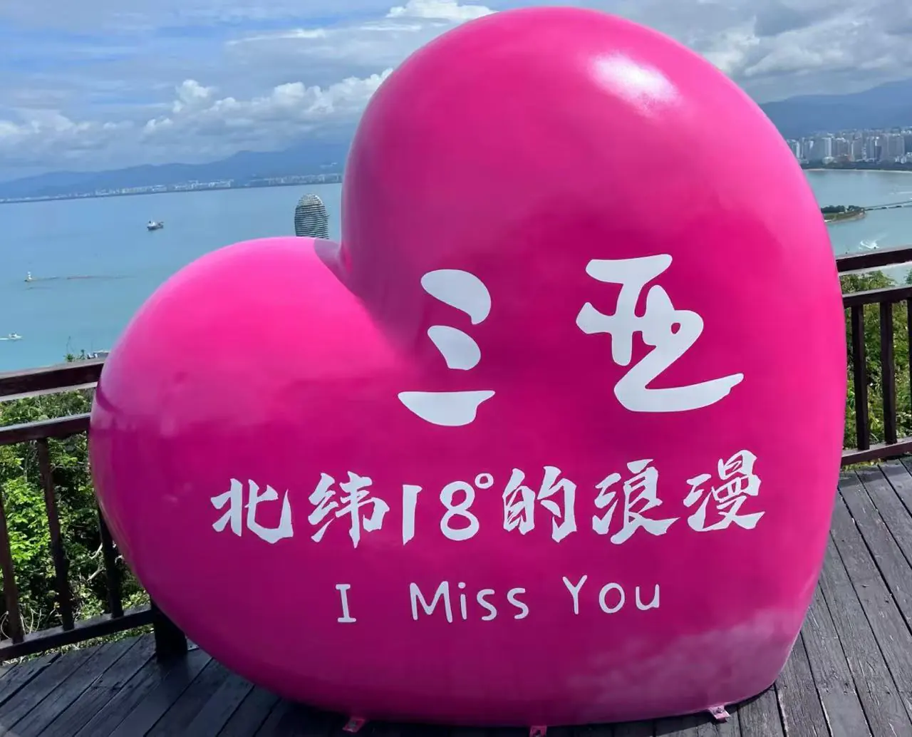 Large pink heart sculpture with white Chinese characters and the English phrase “I Miss You” overlooking a coastal boardwalk.
