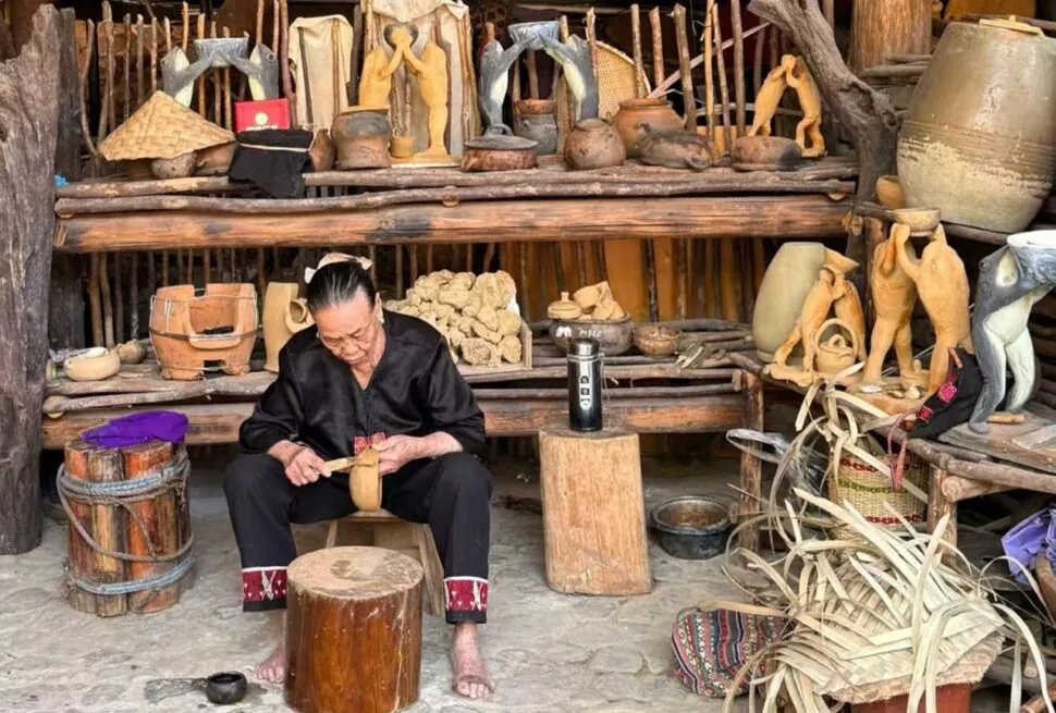 An elderly artisan carving a wooden figure in a rustic workshop filled with pottery and wooden sculptures on shelves.