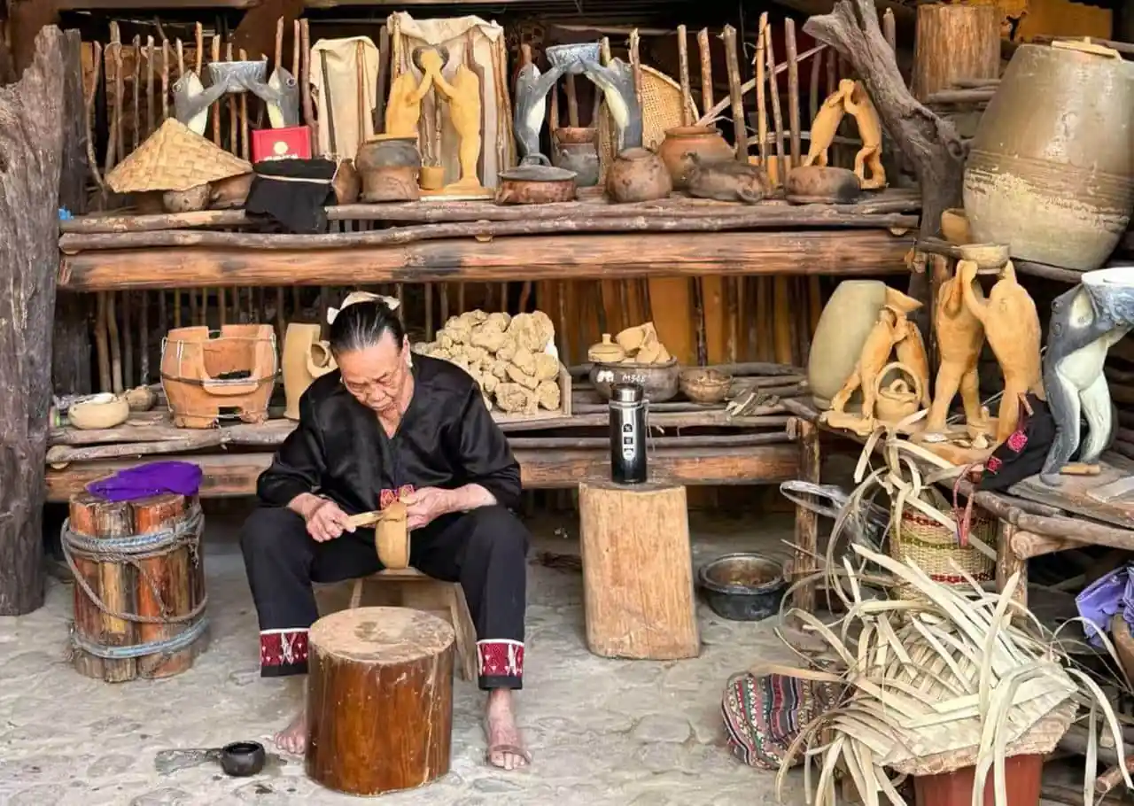 Betel Nut Valley Binglanggu Li and Miao Culture,Sanya,Chinatour An elderly artisan carving a wooden figure in a rustic workshop filled with pottery and wooden sculptures on shelves.
