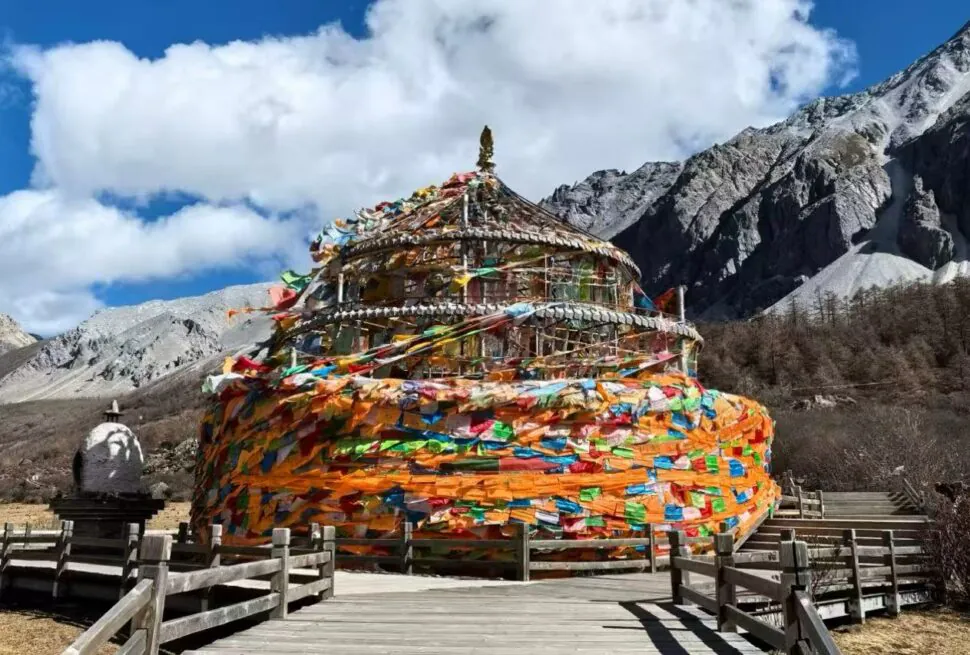 Colorful multi-tiered prayer wheel surrounded by wooden walkway against a rocky mountain landscape and bright blue sky with clouds