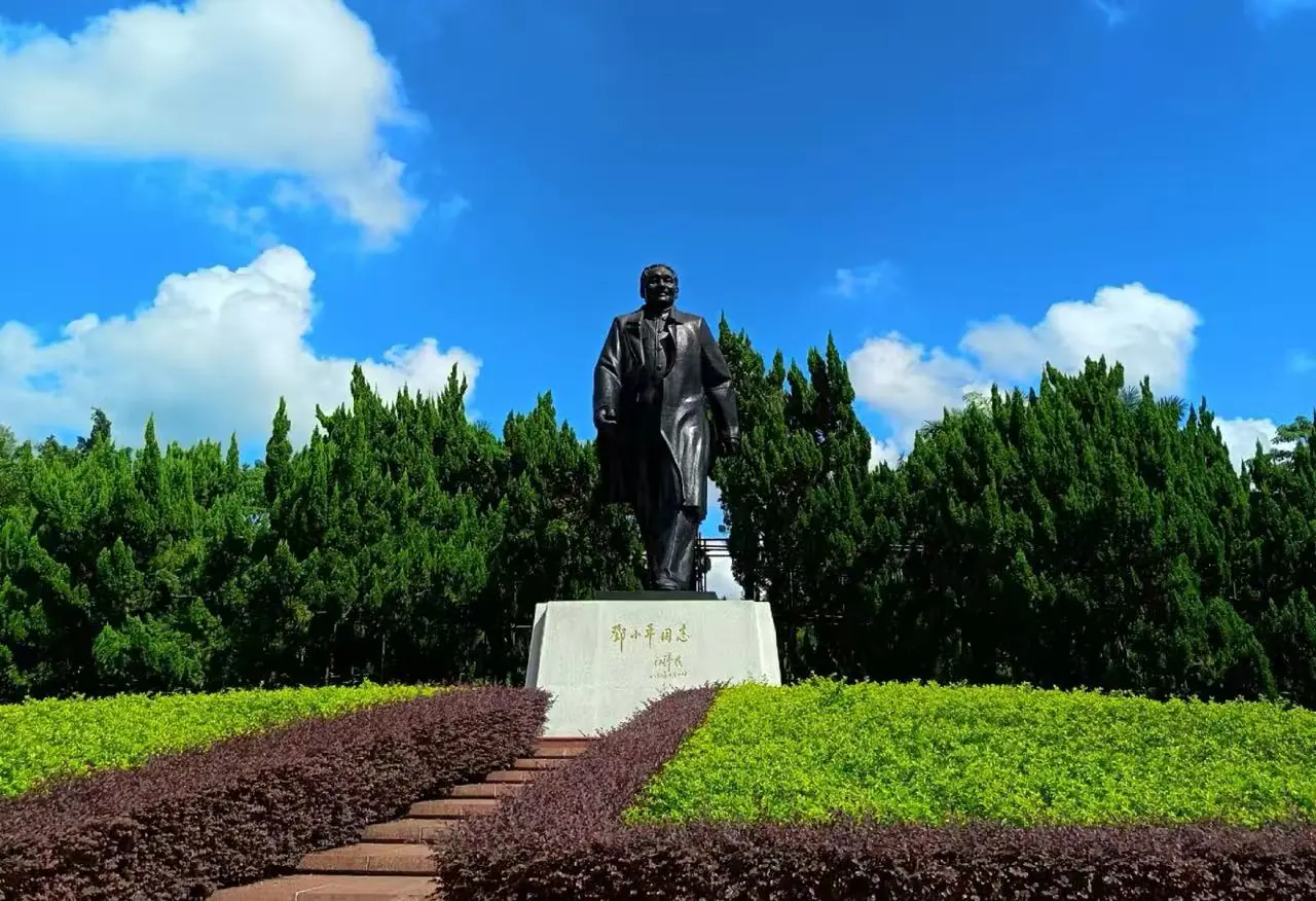 Bronze statue of a man in a suit standing on a pedestal in a landscaped park with shrubs and steps.