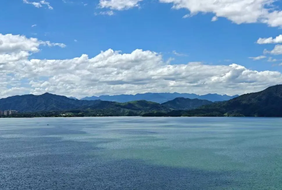 Taiping Lake,Huangshan,Chinatour Calm turquoise lake with distant green mountains under a bright blue sky and fluffy white clouds; a small town sits along the shoreline on the left.