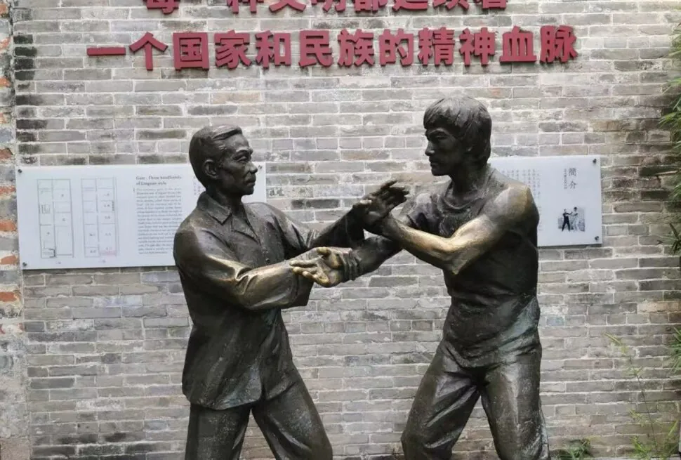 Bruce Lee’s Ancestral Home,Foshan,Chinatour Bronze statue of two men sparring in martial arts in front of a brick wall with Chinese inscription.
