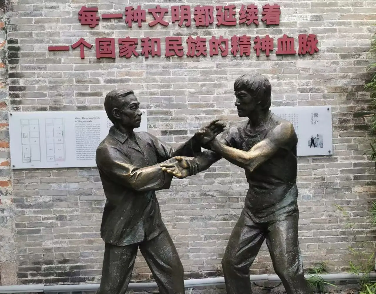 Bruce Lee’s Ancestral Home,Foshan,Chinatour Bronze statue of two men sparring in martial arts in front of a brick wall with Chinese inscription.