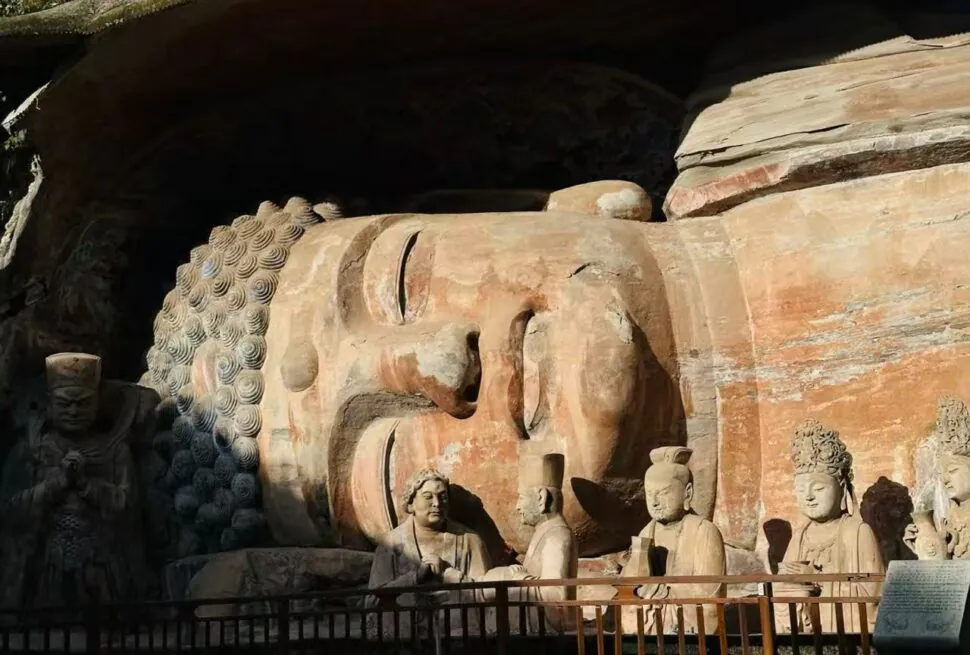 Large reclining Buddha carved in rock inside a cave, with several standing and seated stone figures nearby and a railing in front.