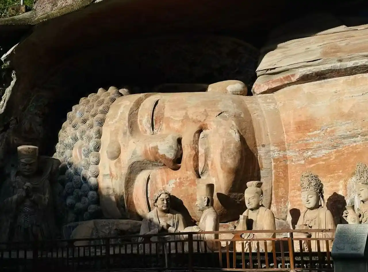 Dazu Rock Carvings Buddhist Sites,Chongqing,Chinatour Large reclining Buddha carved in rock inside a cave, with several standing and seated stone figures nearby and a railing in front.