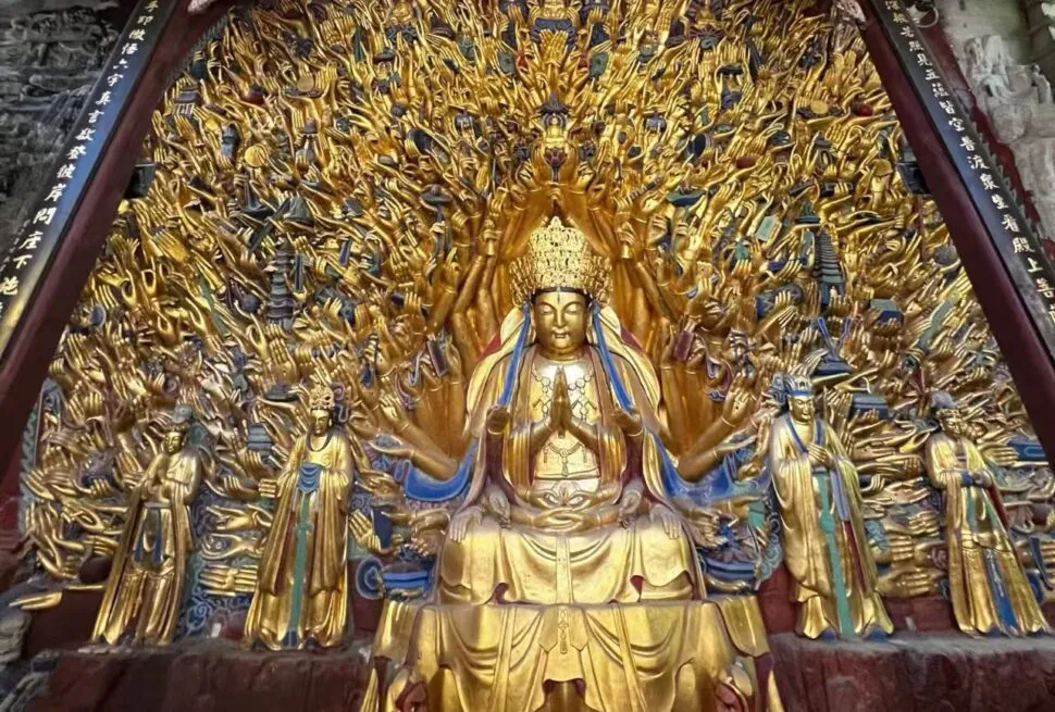 Golden statue of a multi-armed bodhisattva seated in meditation, surrounded by many outstretched arms and smaller figures in a temple setting.