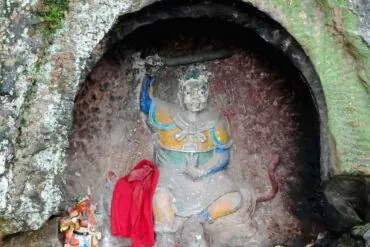 Statue of a seated deity carved into a rocky cave, with yellow and blue clothing, a red cloth draped nearby, and small figurines around the altar in a moss-covered alcove.