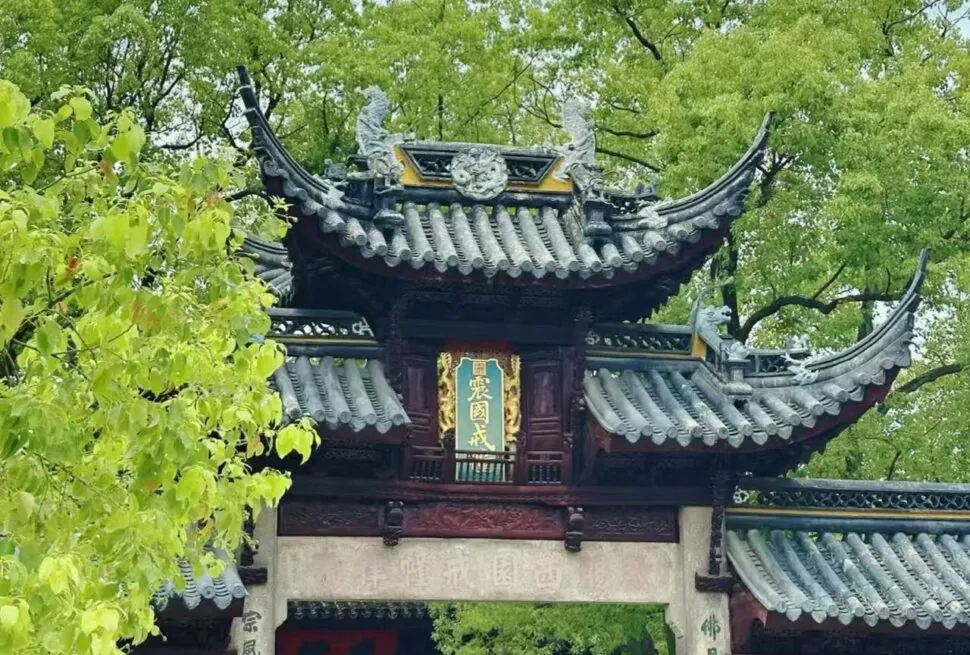 Traditional Chinese temple gate with curved eaves and ornate roof, green signboard with gold characters, surrounded by trees.