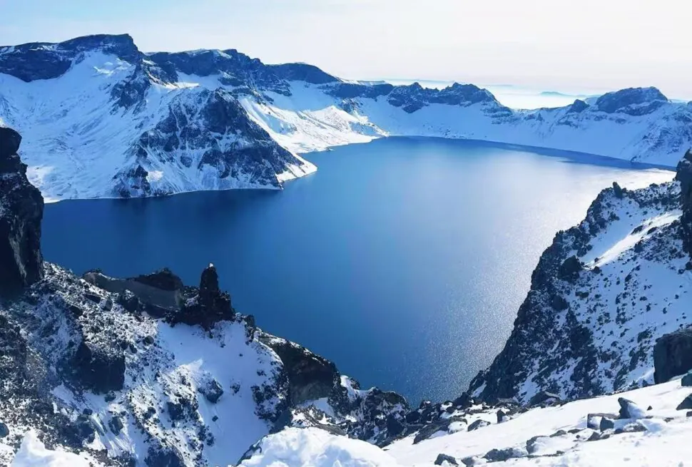 Snow-covered mountains surround a deep blue alpine lake under a pale sky, rocky ridges in the foreground.