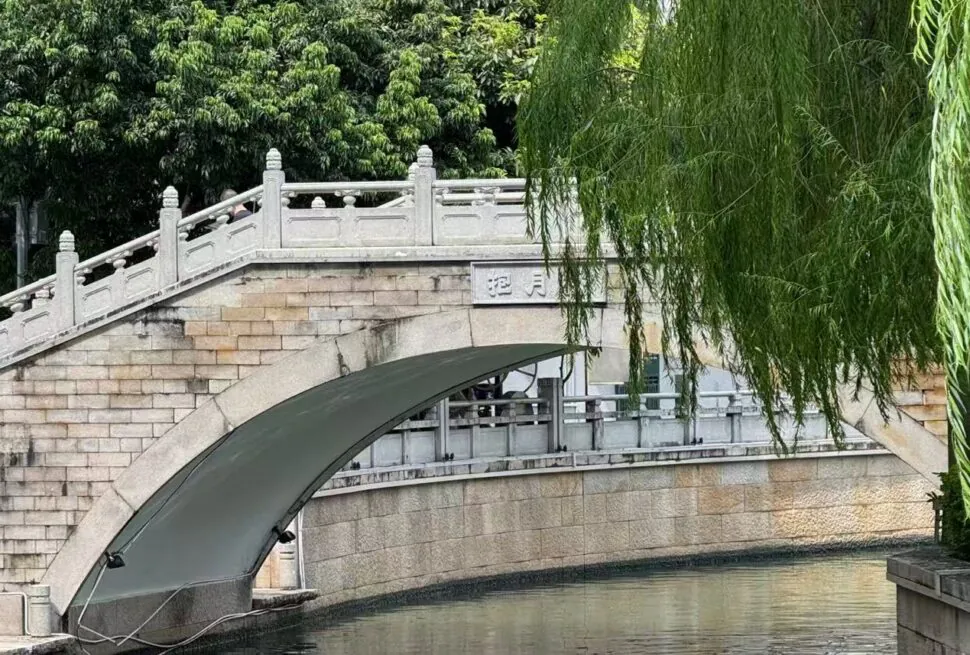 Stone arched bridge over a narrow canal, with a sloped stone ramp and hanging willow branches on the right; lush trees in the background.