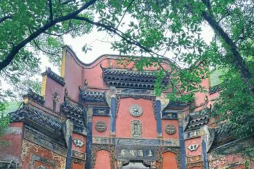 Ornate red Chinese temple gate with blue-gray decorative panels and gold carvings, framed by green trees.