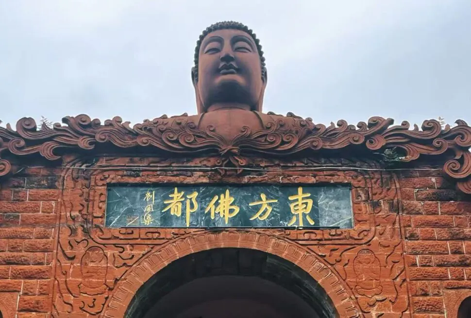 Buddha head statue perched above an ornate red brick gateway with gold Chinese inscription plaque.